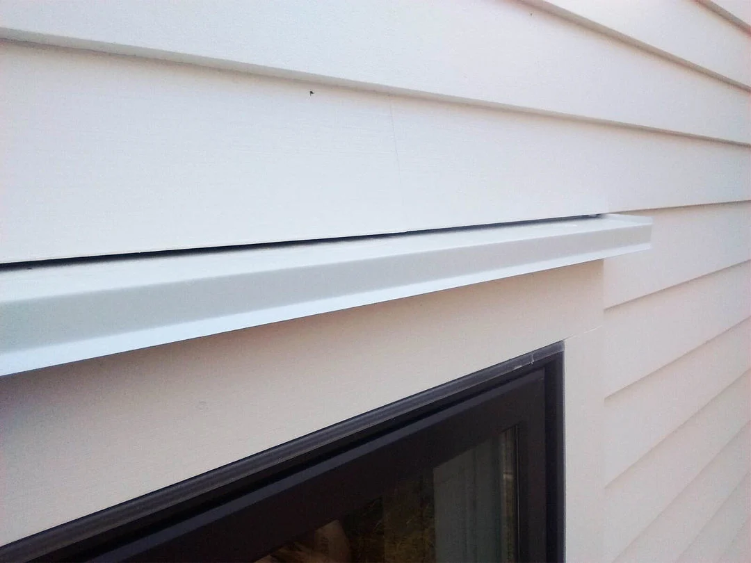A close-up of the top-right corner of a black window with white window framing in a home with horizontal panel siding, showing a white drip cap that’s been installed to better shield and protect the window from water.