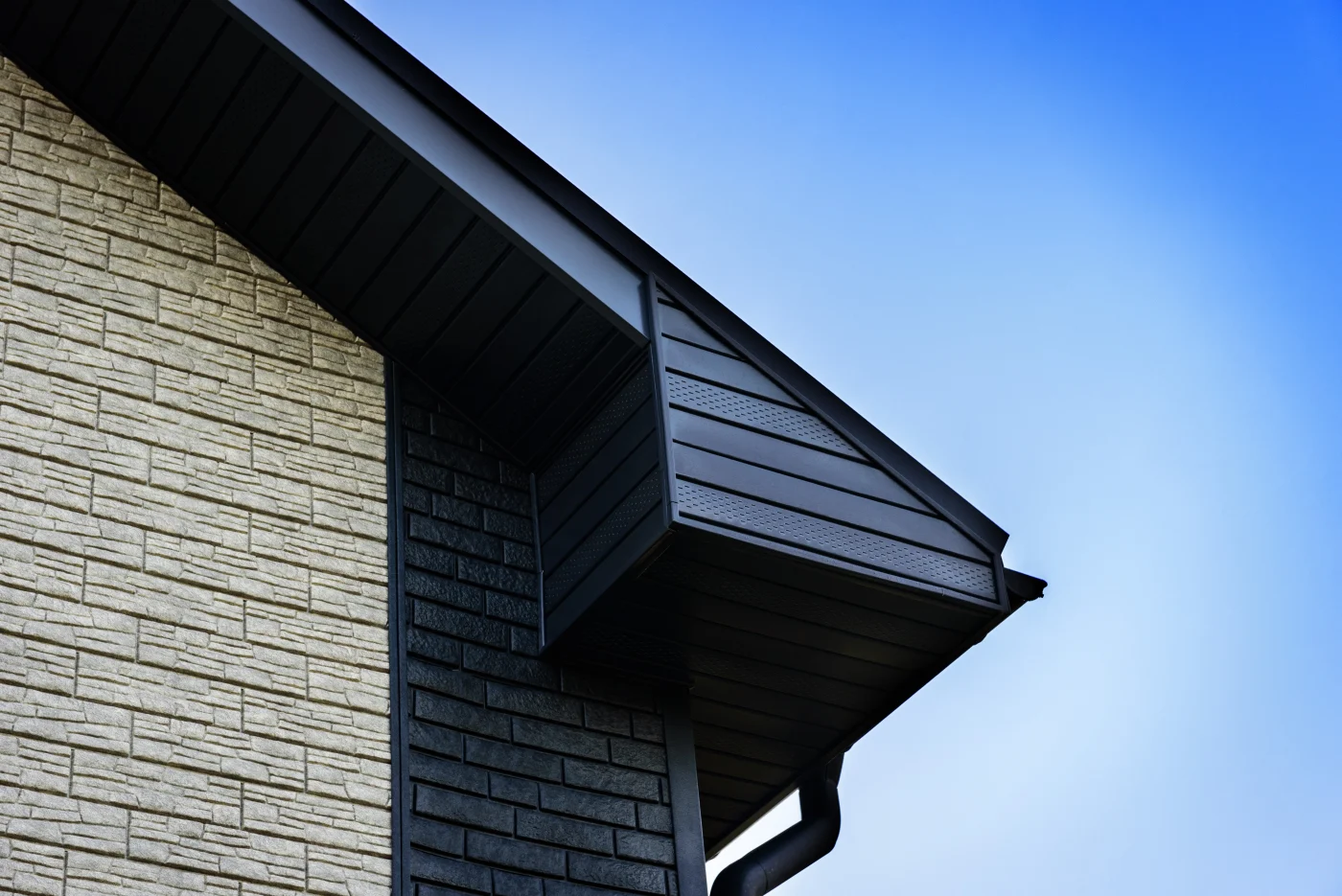 Additional Exterior Remodeling Services For Colorado Springs Homeowners A close-up on the corner of a home’s roof shows the dark-colored soffit, fascia, gutter system, and different kinds of light and dark brick veneer siding up against a blue sky background.