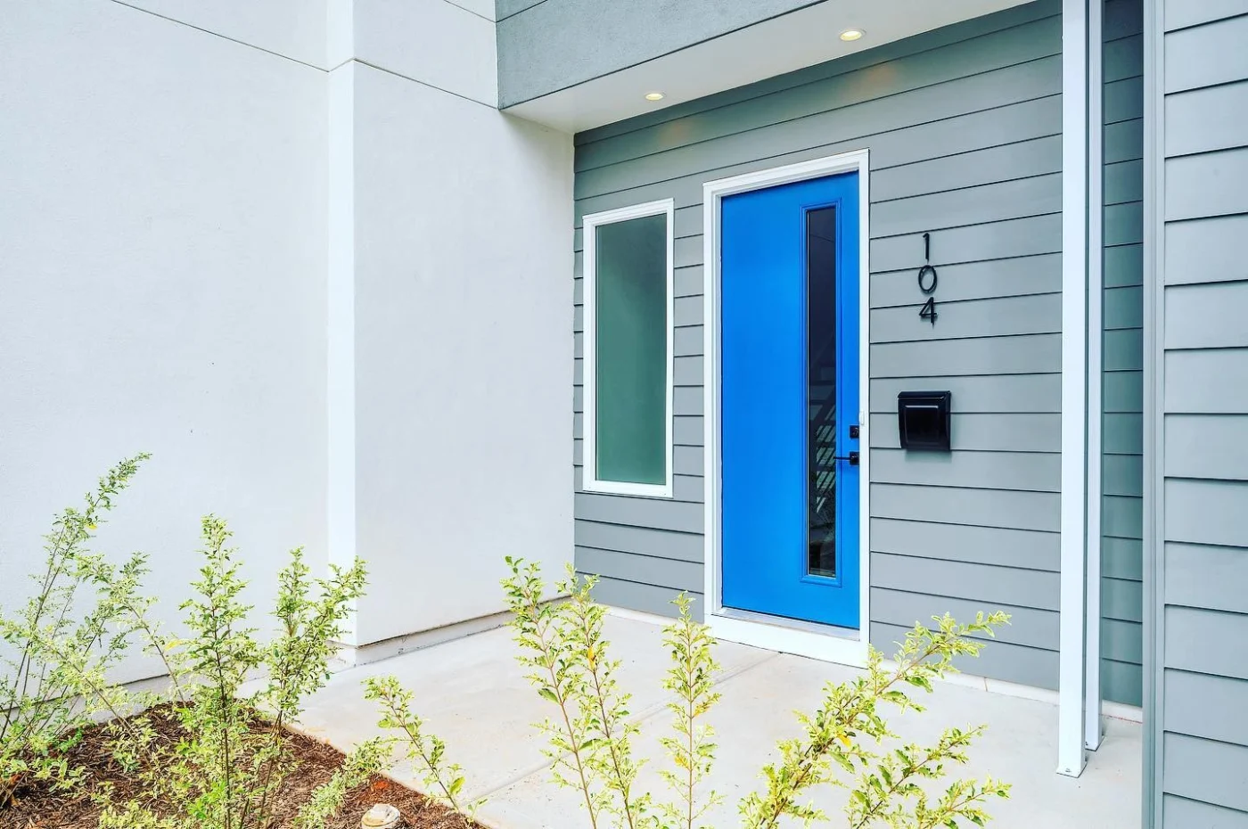 A small front entryway to a condo with a small black mailbox up against light gray horizontal panel siding. There’s also a large picture window with privacy glass next to a bright blue Therma-Tru single-entry door with a thin slit of clear glass going down it and matte black hardware.