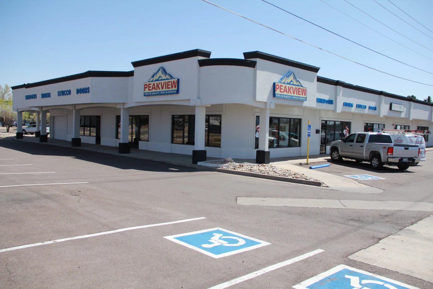 A pictures of both sides of the large Peakview Windows, Siding & Stucco exterior home remodeling showroom from the outside on South Academy Boulevard in Colorado Springs, showing the white building with their brand logo and the words “Windows” “Siding” “Stucco” and “Doors” along the wall.