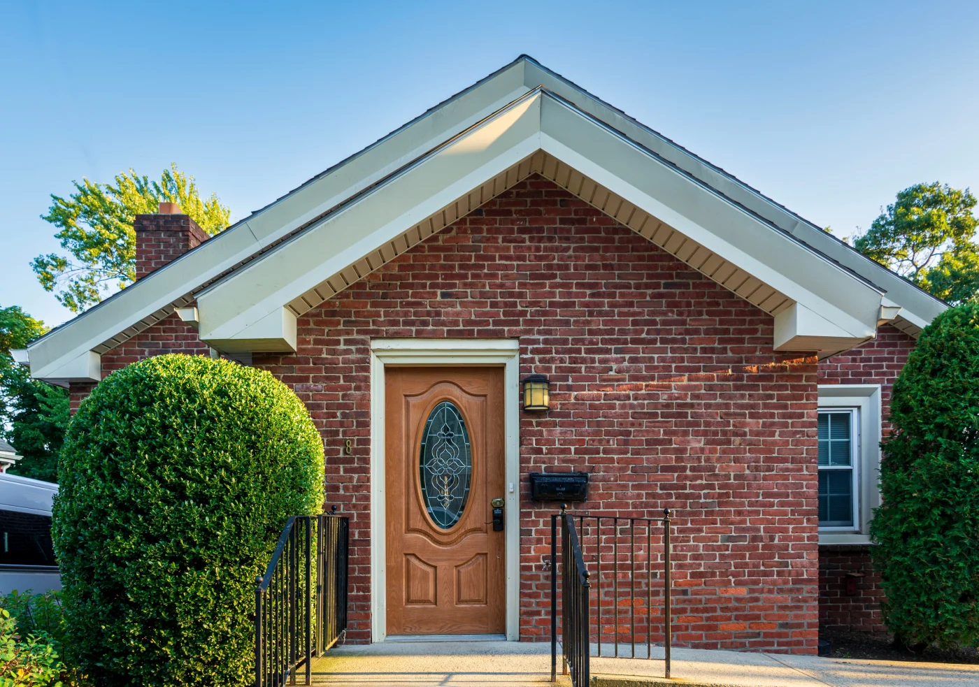 Residential Fascia Services In Colorado Springs The simple front entryway of a red brick single-story home with a wood single-entry door with decorative glass in the middle, and two large sections of white fascia showing that matches the white windows and window/door framing.