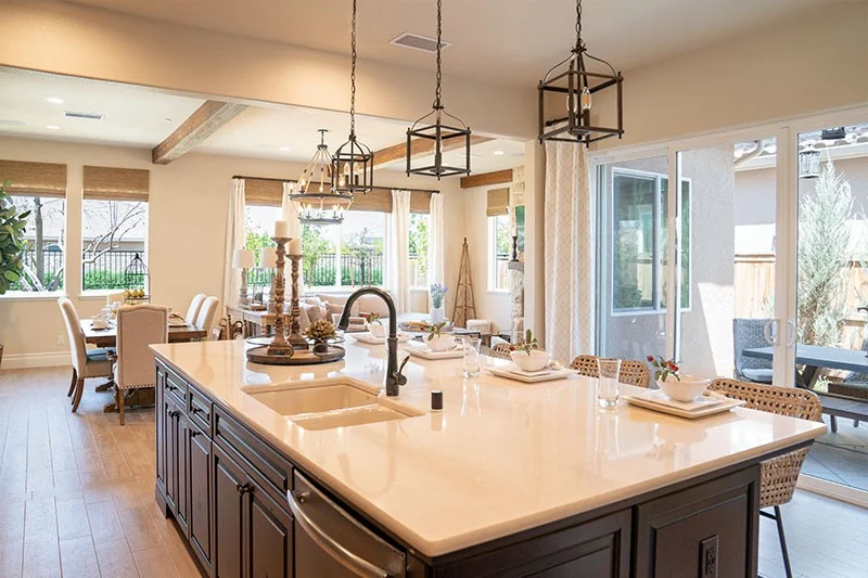 A lovely, well-lit, furnished, open-concept kitchen with a large island, dining table, and lounging area with sets of Anilin vinyl replacement windows from their Del Mar Collection with open blinds and a white sliding glass door to the right.