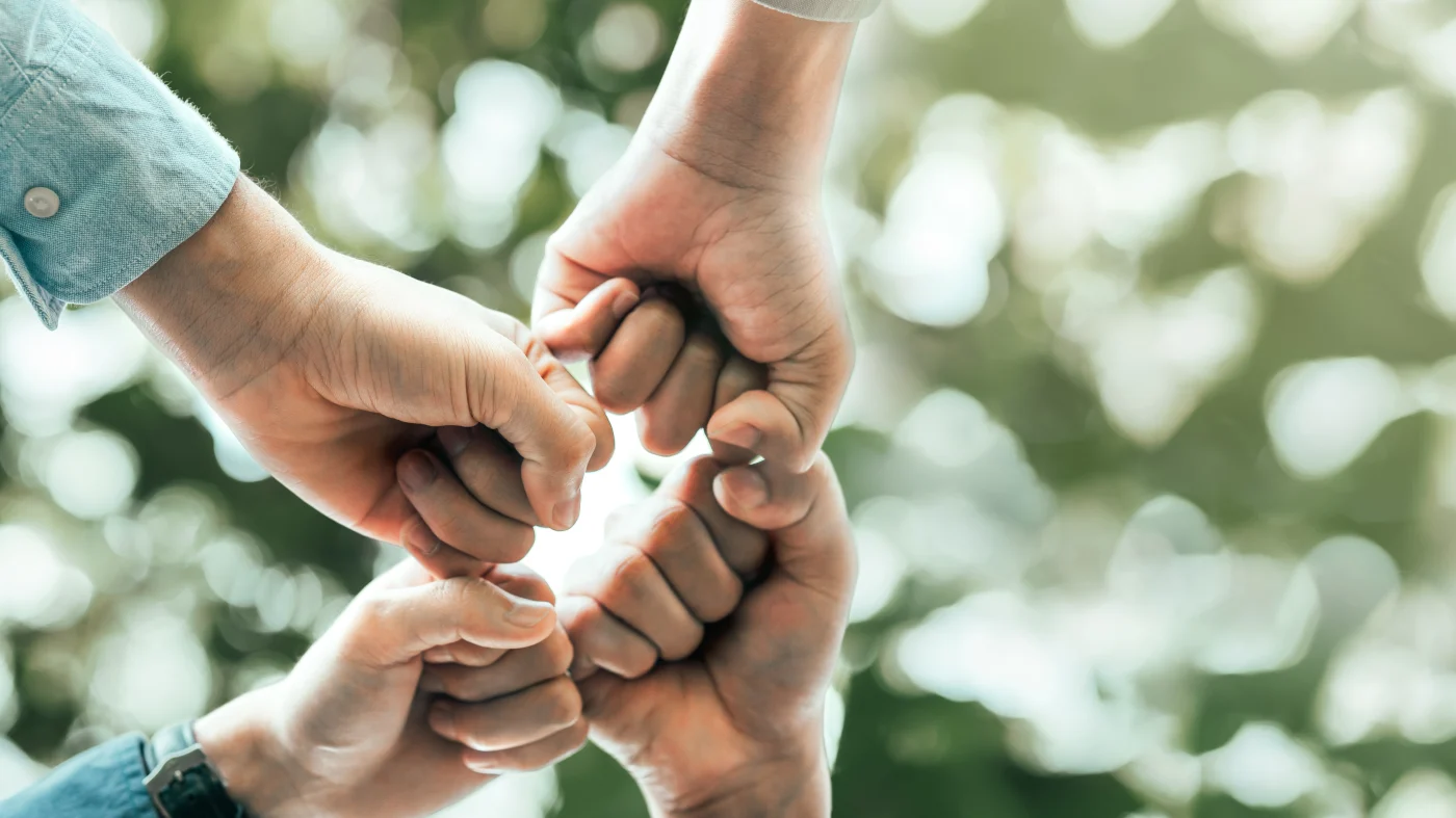 Four hands in fists meeting together, symbolizing family, against a blurred green and white background that resembles blurred plants.