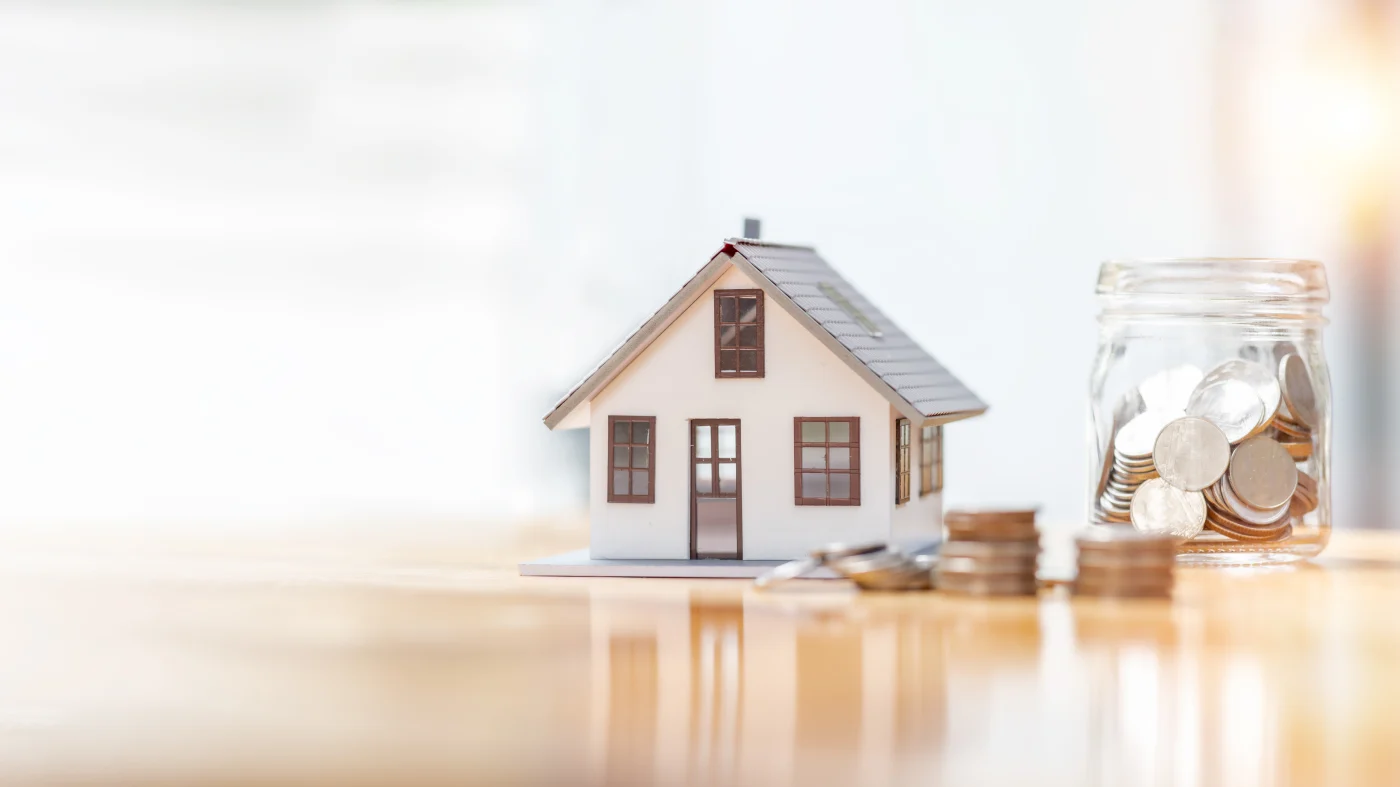 There is a miniature home with a single entry door, two bottom floor windows and an attic window sitting on a clear polished wood table. Beside the small home are a few little piles of coins next to a small glass jar with more coins, symbolizing financing an exterior home remodeling project.