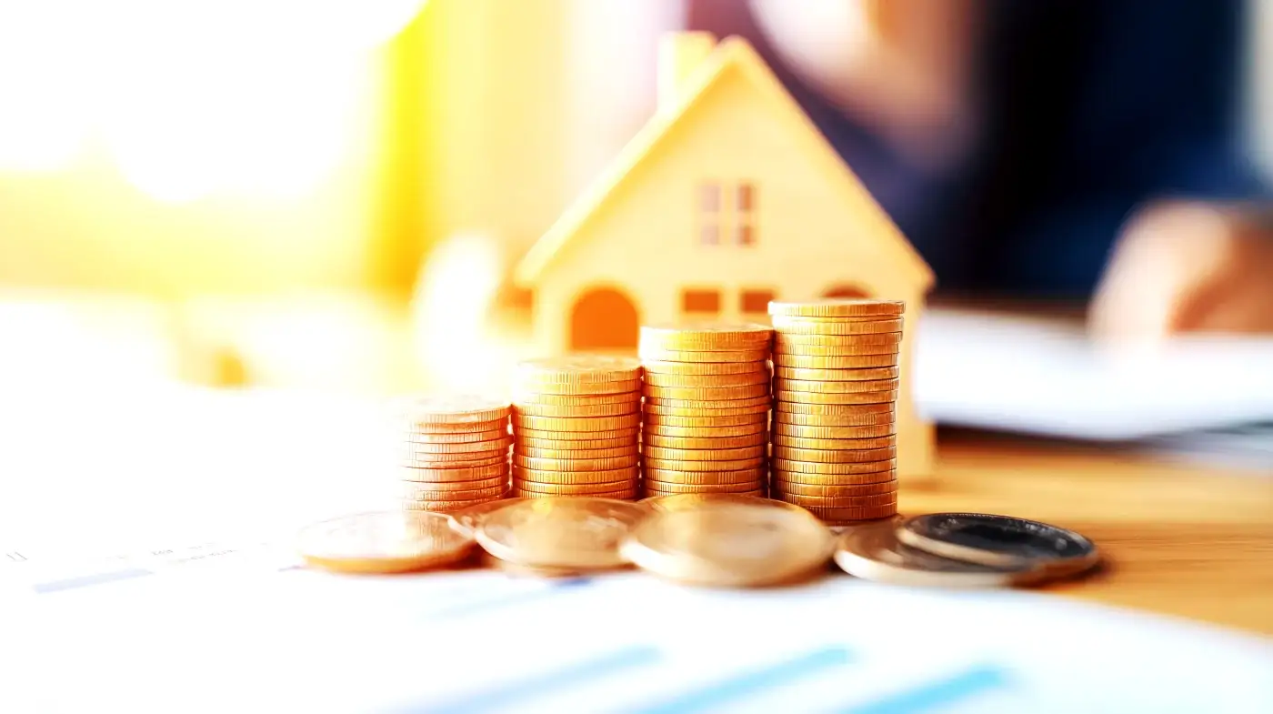 Financing Residential Exterior Remodeling Projects In The Colorado Springs Region A blurry white and yellow background with a miniature home, while in focus are four stacks of coins with more coins scattered around them, symbolizing financing a home exterior remodeling project