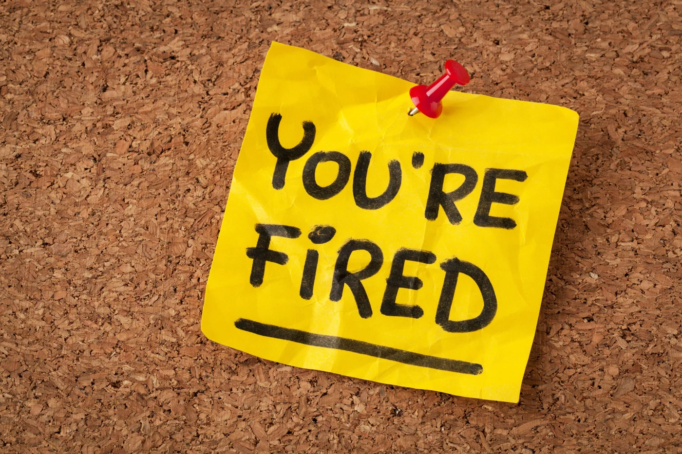 There’s a yellow sticky note tacked to a pinboard with a red tack that says “You’re Fired” with the word “fired” underlined.