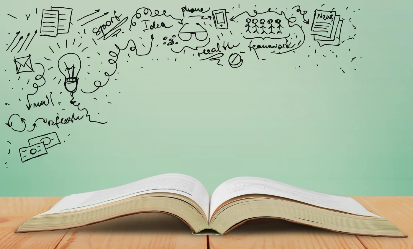 A book is sitting open on a wooden desk with a very light green background. There’s a variety of doodles to the upper left, down the left side, and across the top with a lightbulb, glasses, little people, papers, arrows, etc.