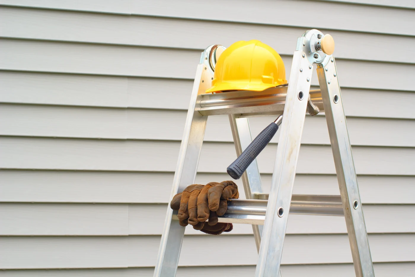 There’s a ladder witting in front of a residential wall with premium horizontal vinyl panel siding, a hard hat wits on the top of the ladder, a hammer hangs from the top step, and there are gloves resting atop the second step.