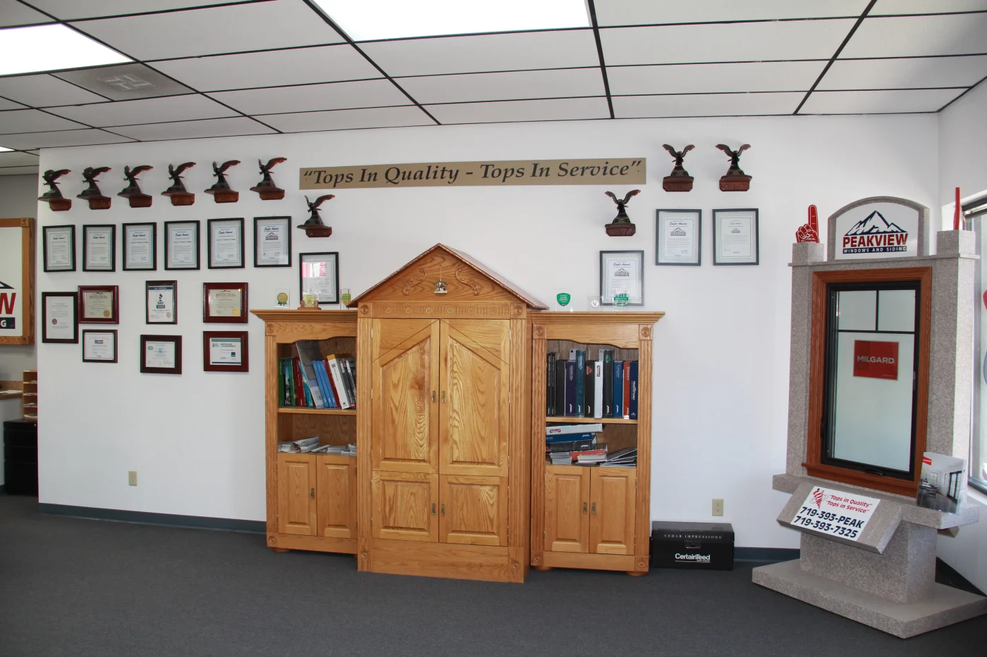 A wall in the Peakview Windows, Siding & Stucco exterior remodeling showroom in Colorado Springs, showing ten local Eagle Awards for customer service along with different plaques, licenses, and certificates, as well as a lovely golden hutch with folders and books, and a window display to the right.