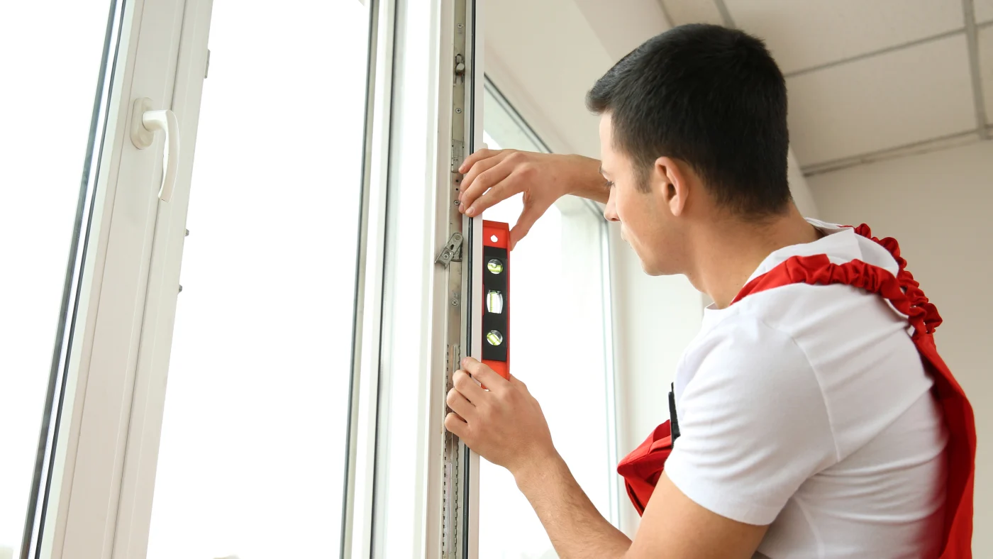 A specialized window installer wearing a white t-shirt and overalls using a level on a newly-installed replacement window in white.