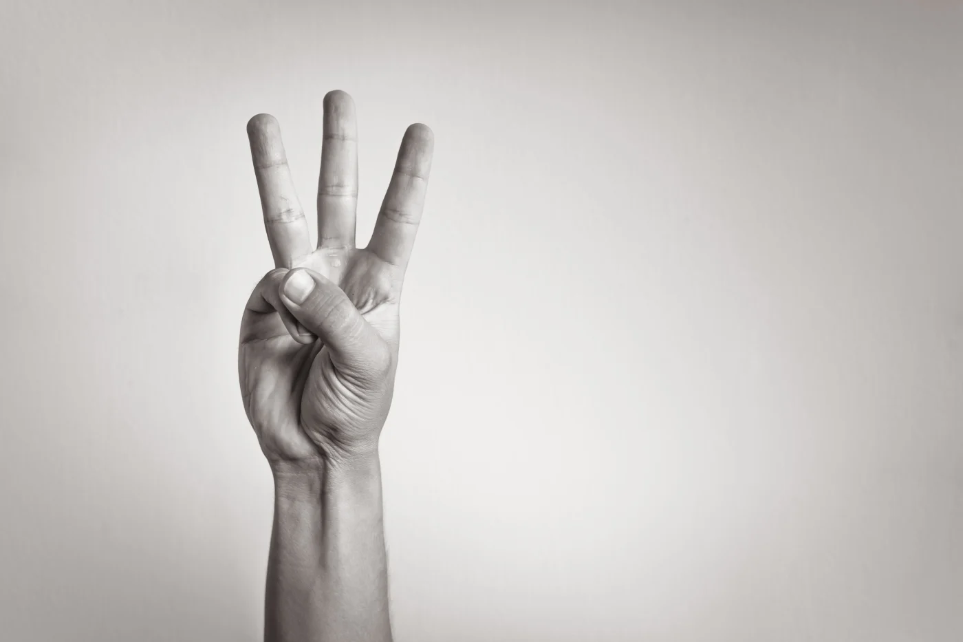 A black and white photo showing a hand in the air with three fingers pointing up symbolizing the number three, with a light gray and white background.