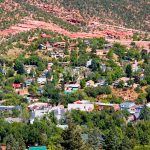 This is a drone shot looking down on a neighborhood of homes in the foothills west of Colorado Springs, CO. They’re nestled amidst lush green trees, and then there’s a more rugged, almost desert-like area going up the hills, with layers of rock visible in the hillside.