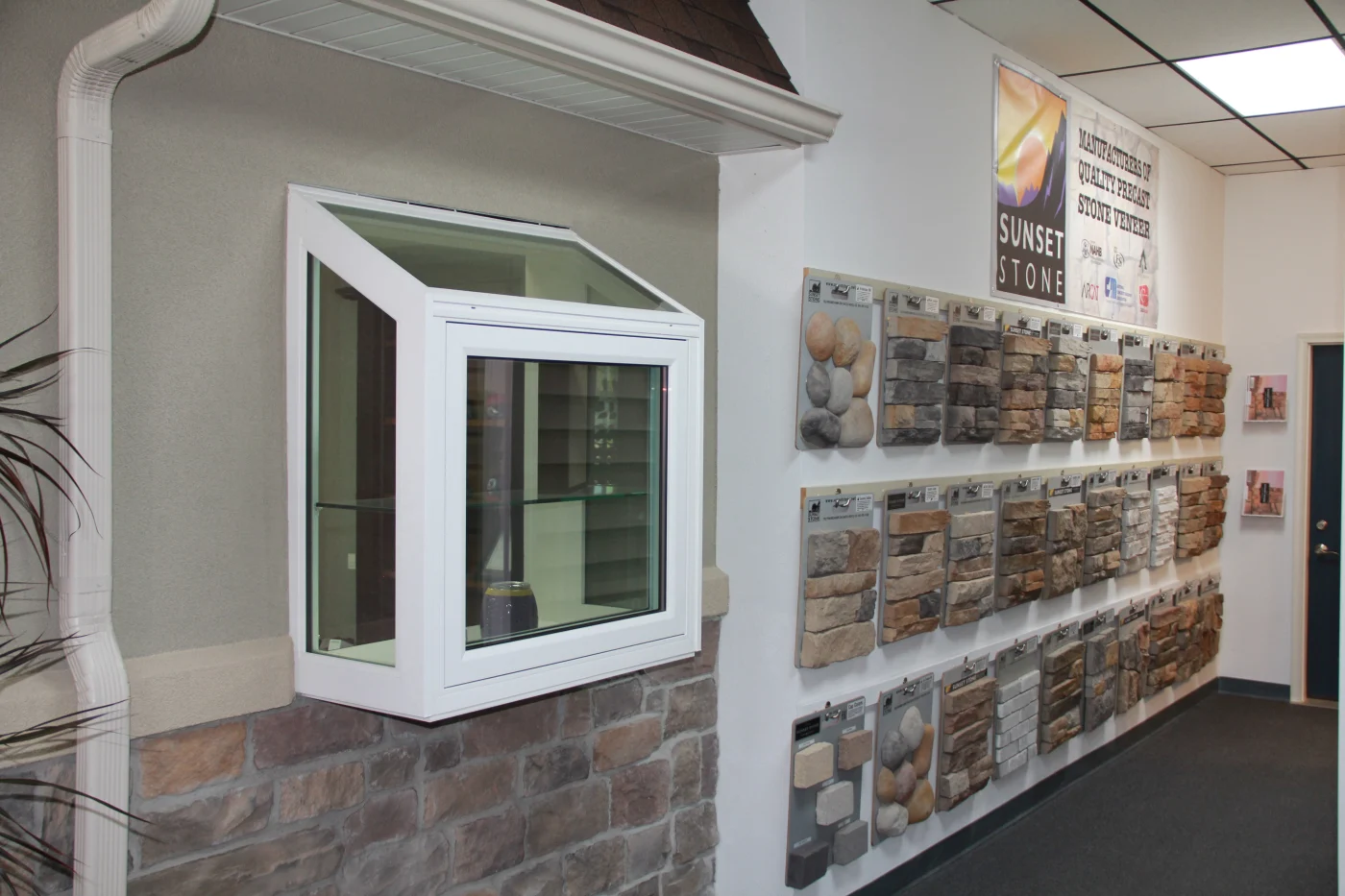 A section of the Peakview Windows, Siding & Stucco showroom in Colorado Springs with a pop-out window in a stucco wall with a stone veneer at the base and a gutter system along with a wall of Sunset Stone veneer sample options to the right.