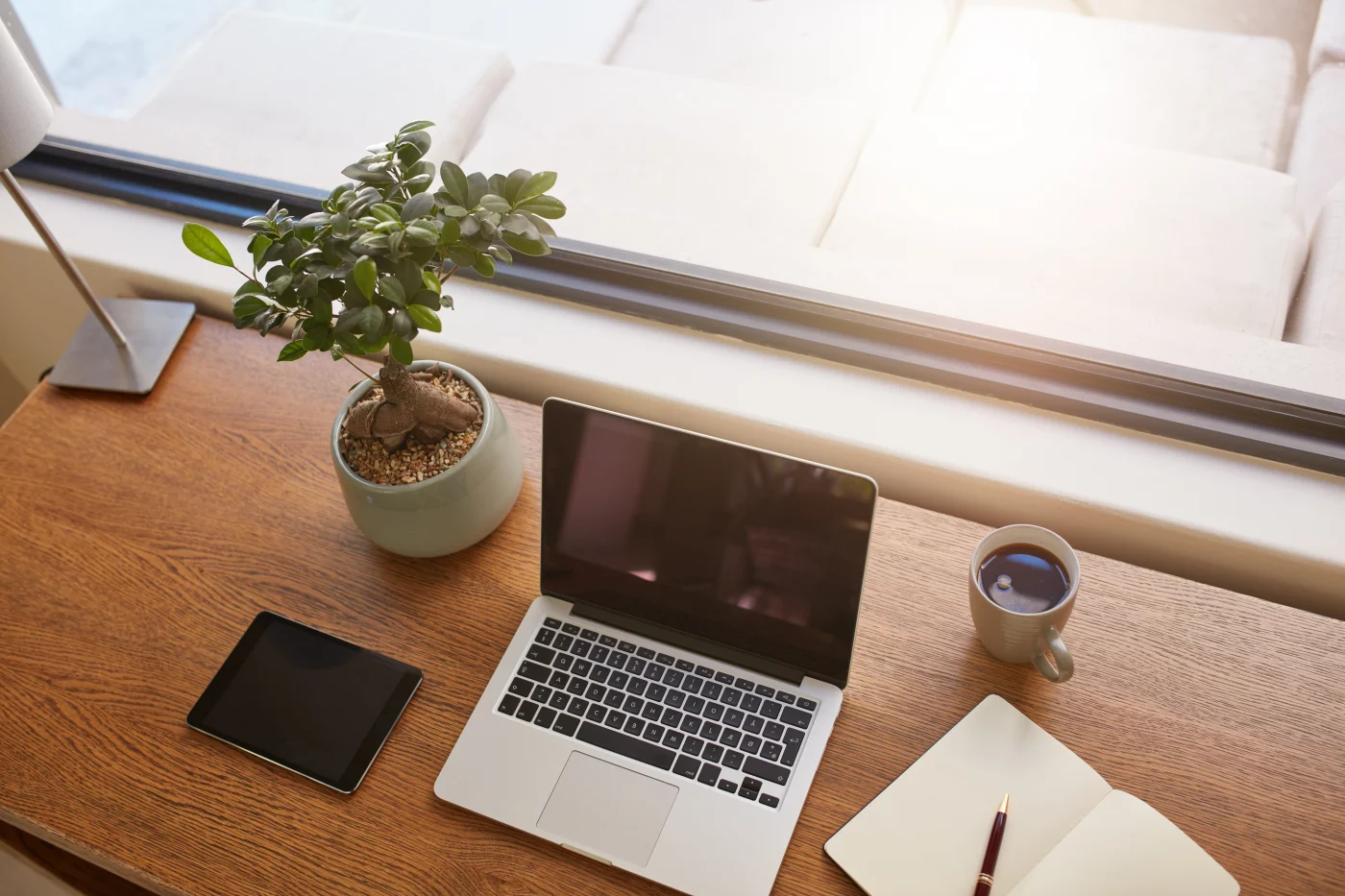 There’s a modern wood desk in a home office with a nice finish, next to and facing a window, featuring an open laptop, a small tablet, a small succulent in a planter, a filled coffee cup, a notepad, a pen, and a small lamp.