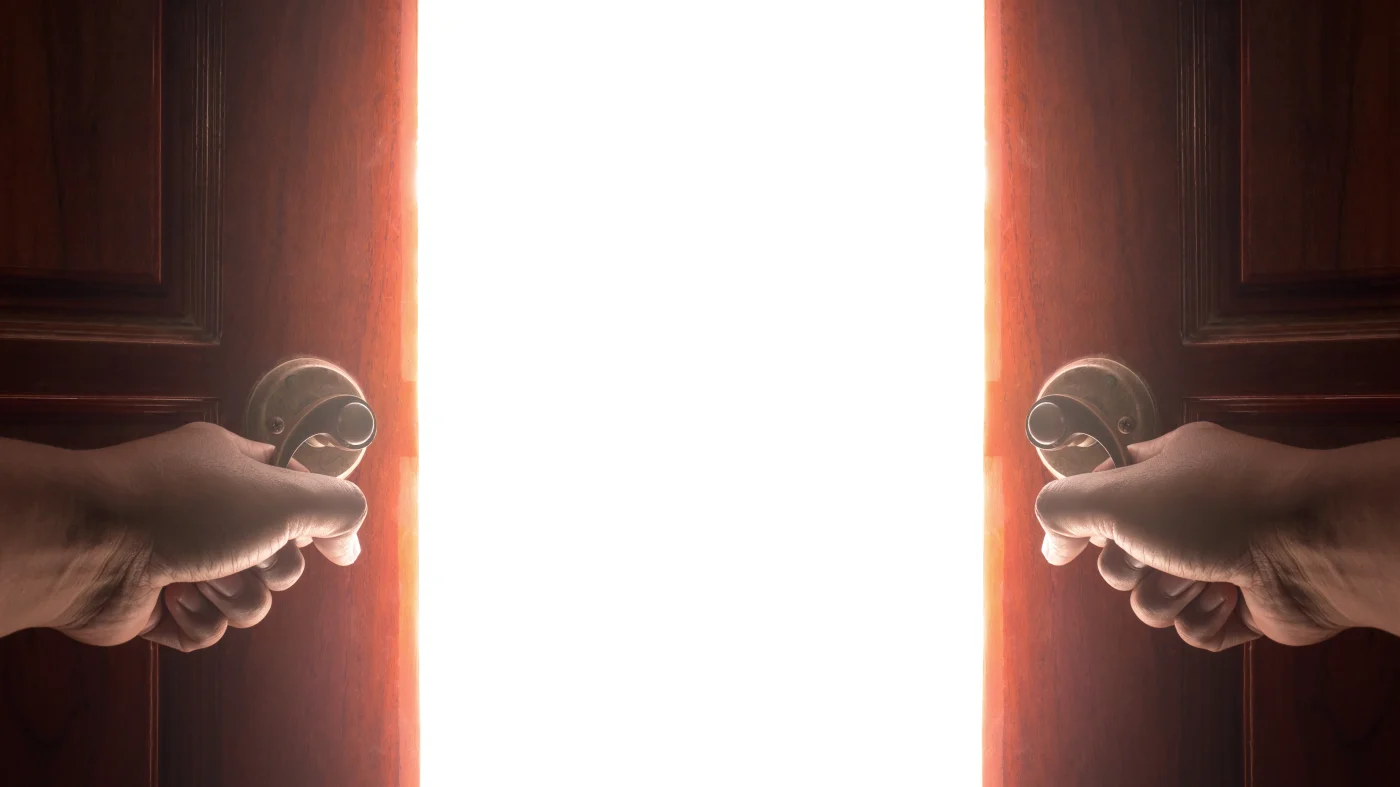 Two hands open a French door to a brightly lit space