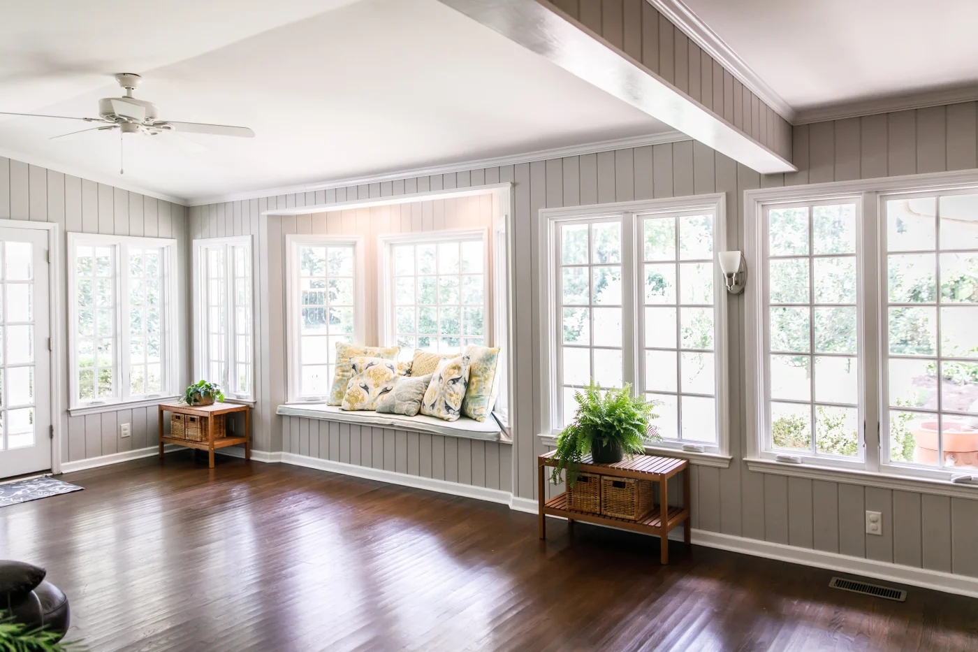 Large And Open Living Room Den Sun Room With Windows On Two Sides And Lots Of Natural Light Flowing In. There Is A Window Seat On One Side And A Leather Couch And Plant On The Other.