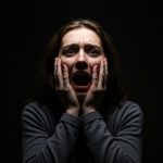 A woman standing against a black background holds her hands against her face while she screams
