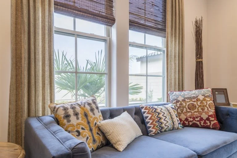 Bright living room with a blue sectional sofa in front of two large energy-efficient windows, framed by beige curtains and bamboo shades
