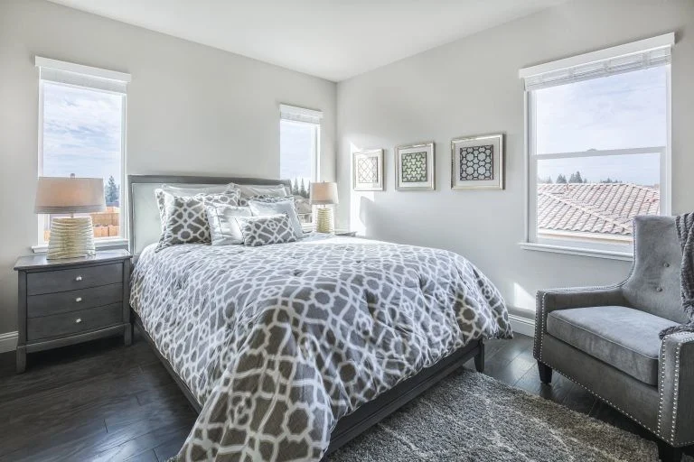 Bright modern bedroom with large energy-efficient windows on two walls, a queen bed with gray patterned bedding, and matching nightstands with lamps.