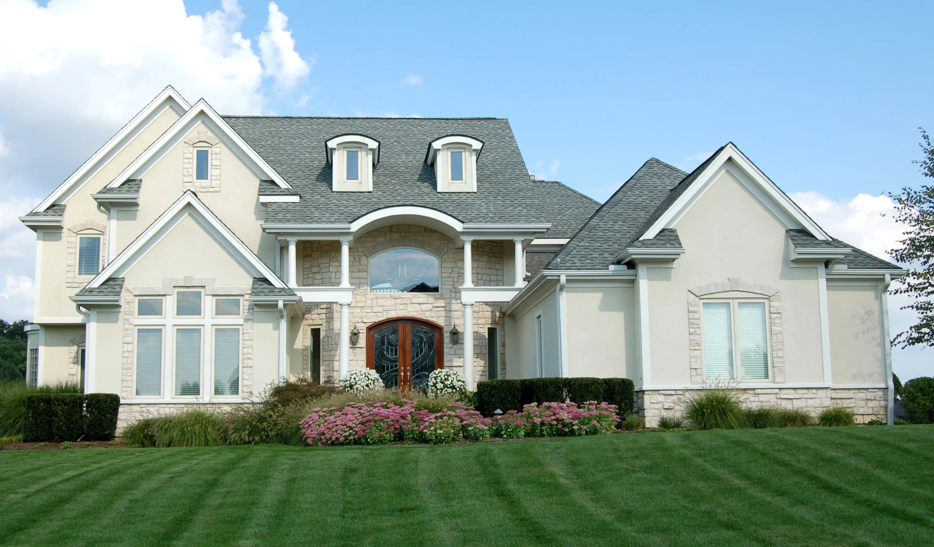 Two-story tan stucco home with stone accent wall, tile roof, landscaped front yard, and wide driveway under a clear blue sky.