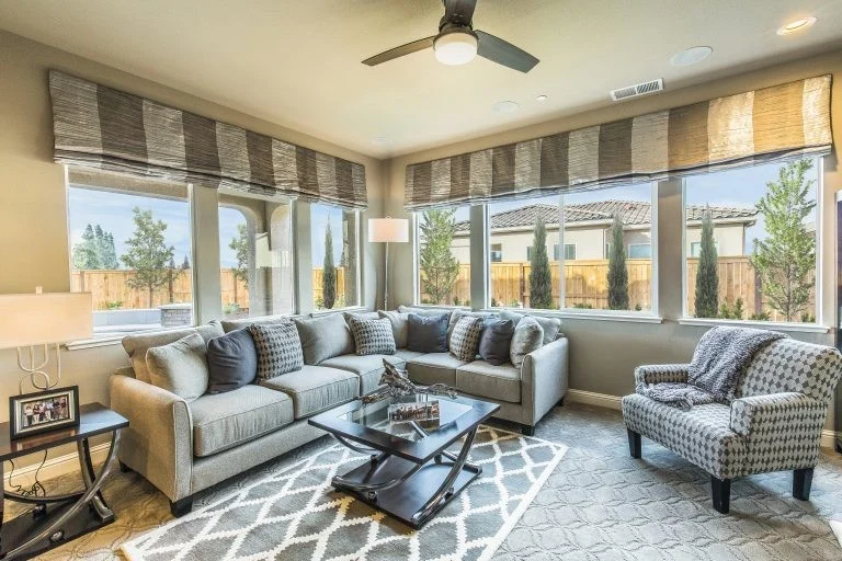 Bright living room with a large gray sectional sofa and houndstooth accent chair, surrounded by tall windows with woven shades.