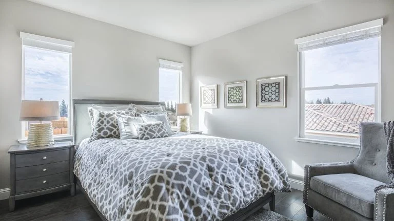 Bright, airy bedroom with a queen bed in gray patterned bedding, two bedside tables with lamps, and three picture frames on the wall.
