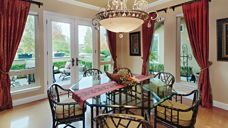 Cozy backyard patio of a modern home at dusk, with French doors and large windows glowing from indoor light, cushioned chairs around a fire table.