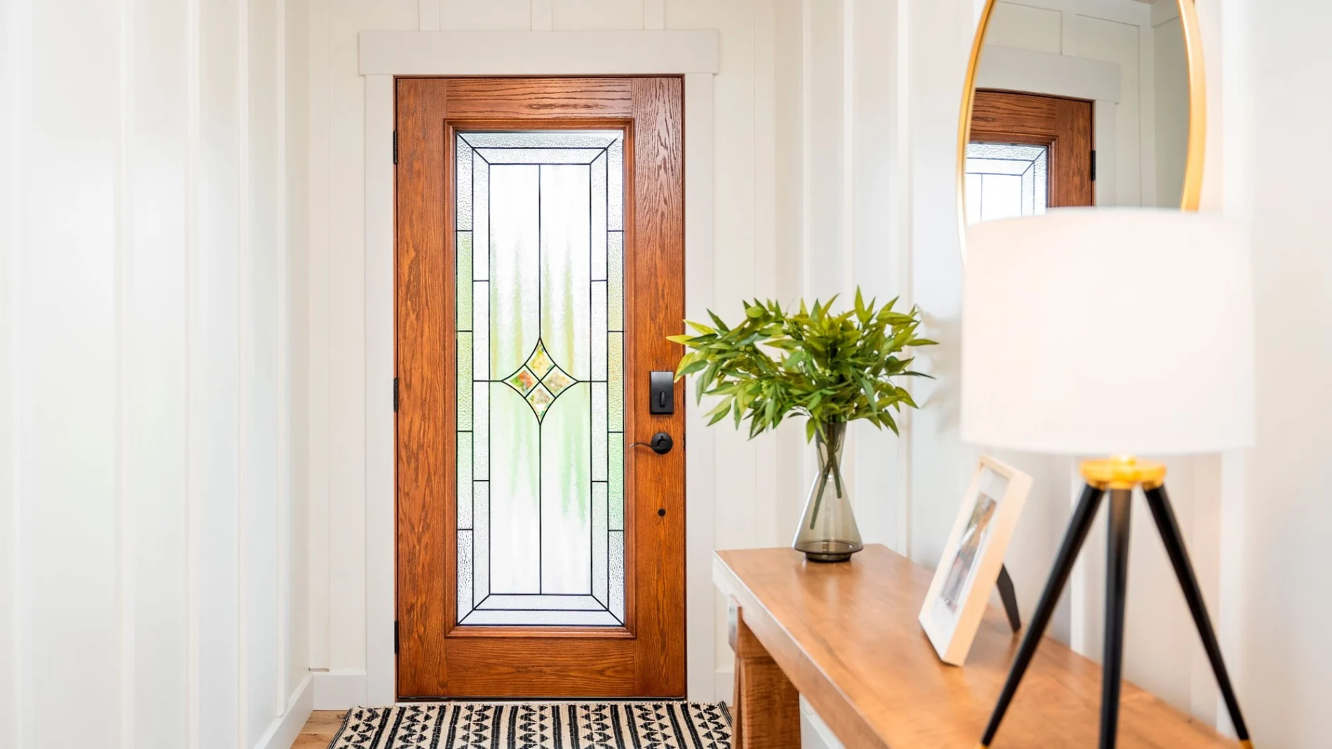 Bright entry hallway featuring a wooden front door with decorative frosted glass panel, flanked by white walls, and a patterned rug.