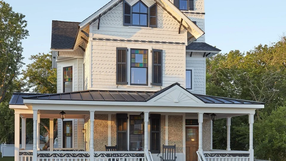 Victorian-style two-story house with white siding, black shutters, colorful stained-glass windows, and a wraparound porch with rocking chairs.