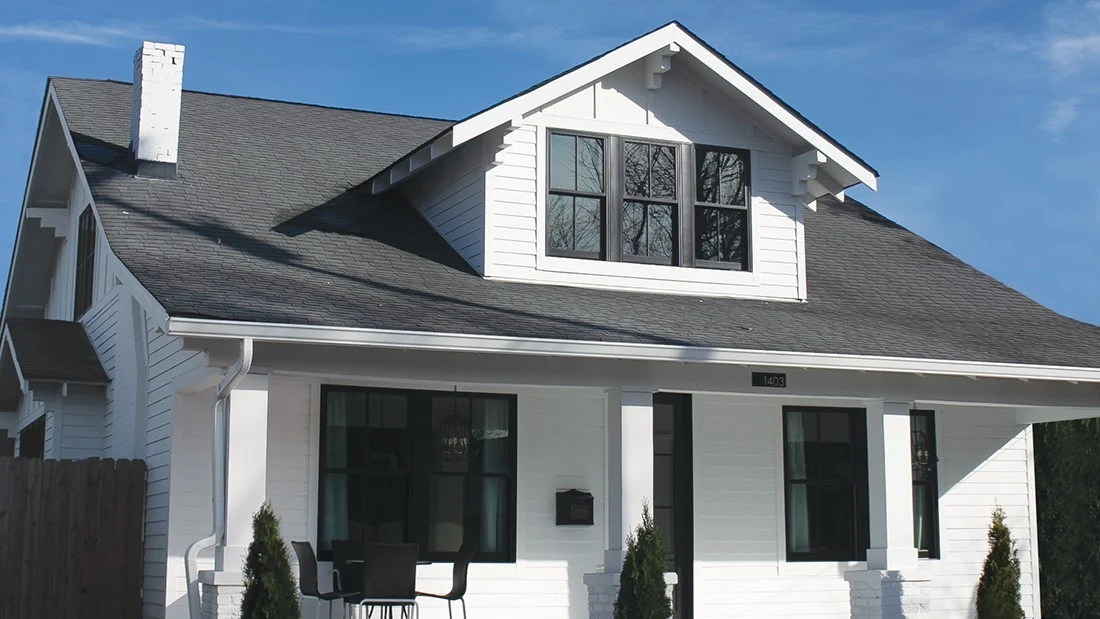 White two-story bungalow with dark shingle roof, black-framed windows, and a small front porch with chairs.