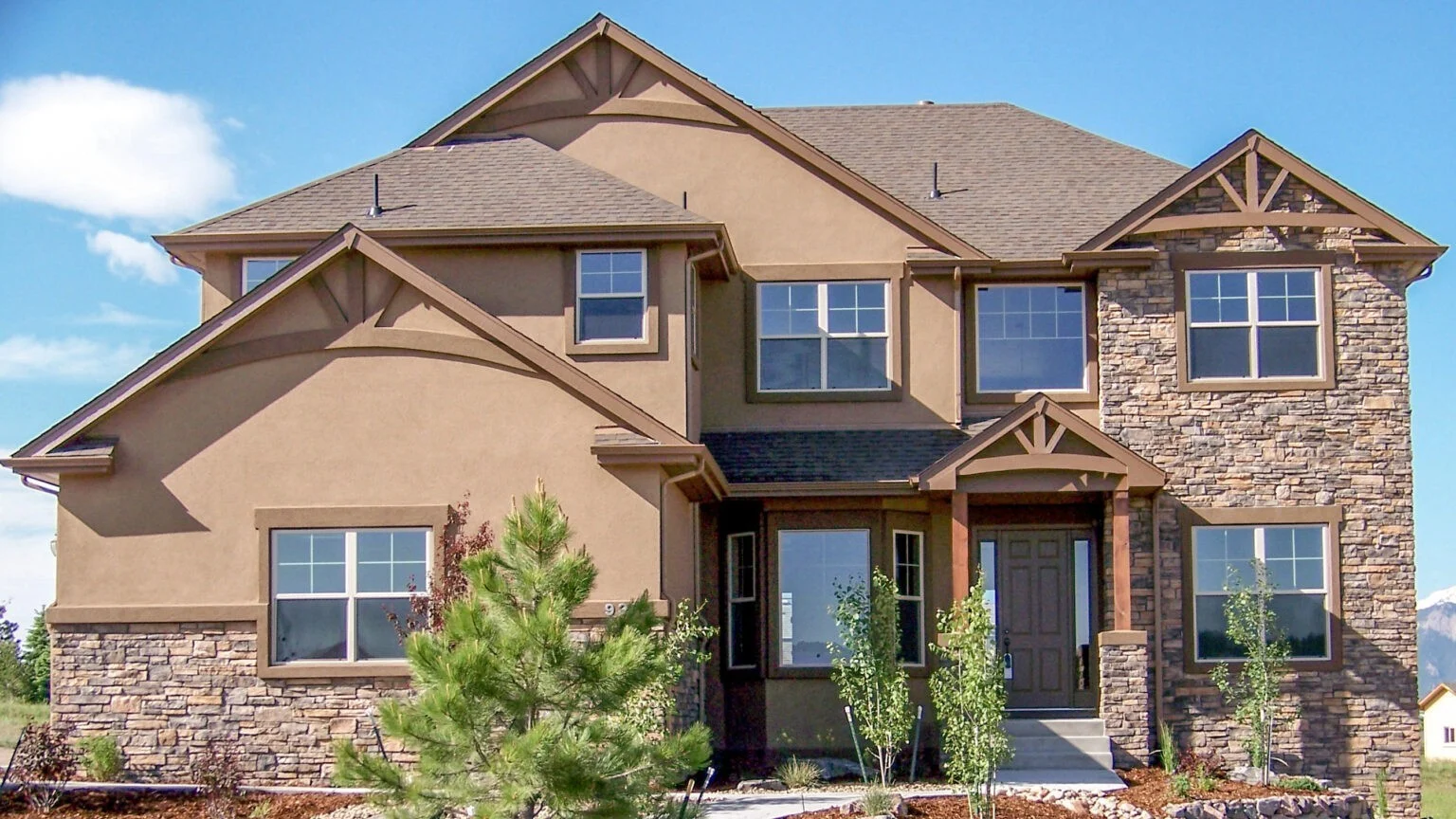Two-story tan stucco home with stone accents and multiple gabled rooflines, featuring a covered front entry, large windows, and young trees and shrubs.