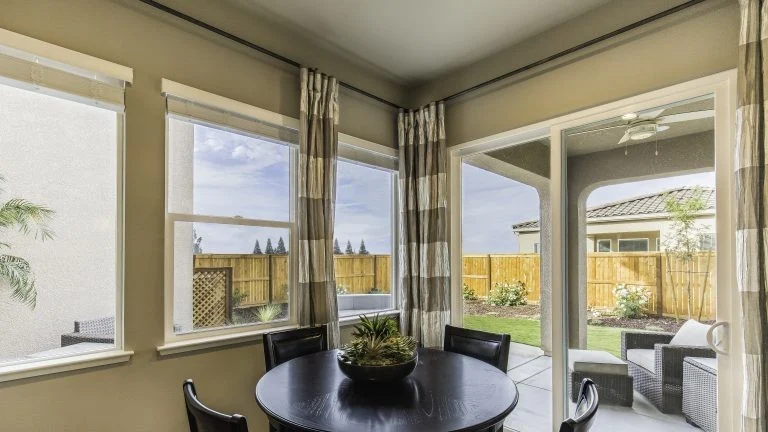 Bright breakfast nook with a round table and four chairs surrounded by large corner windows and a glass door looking out onto a fenced backyard.