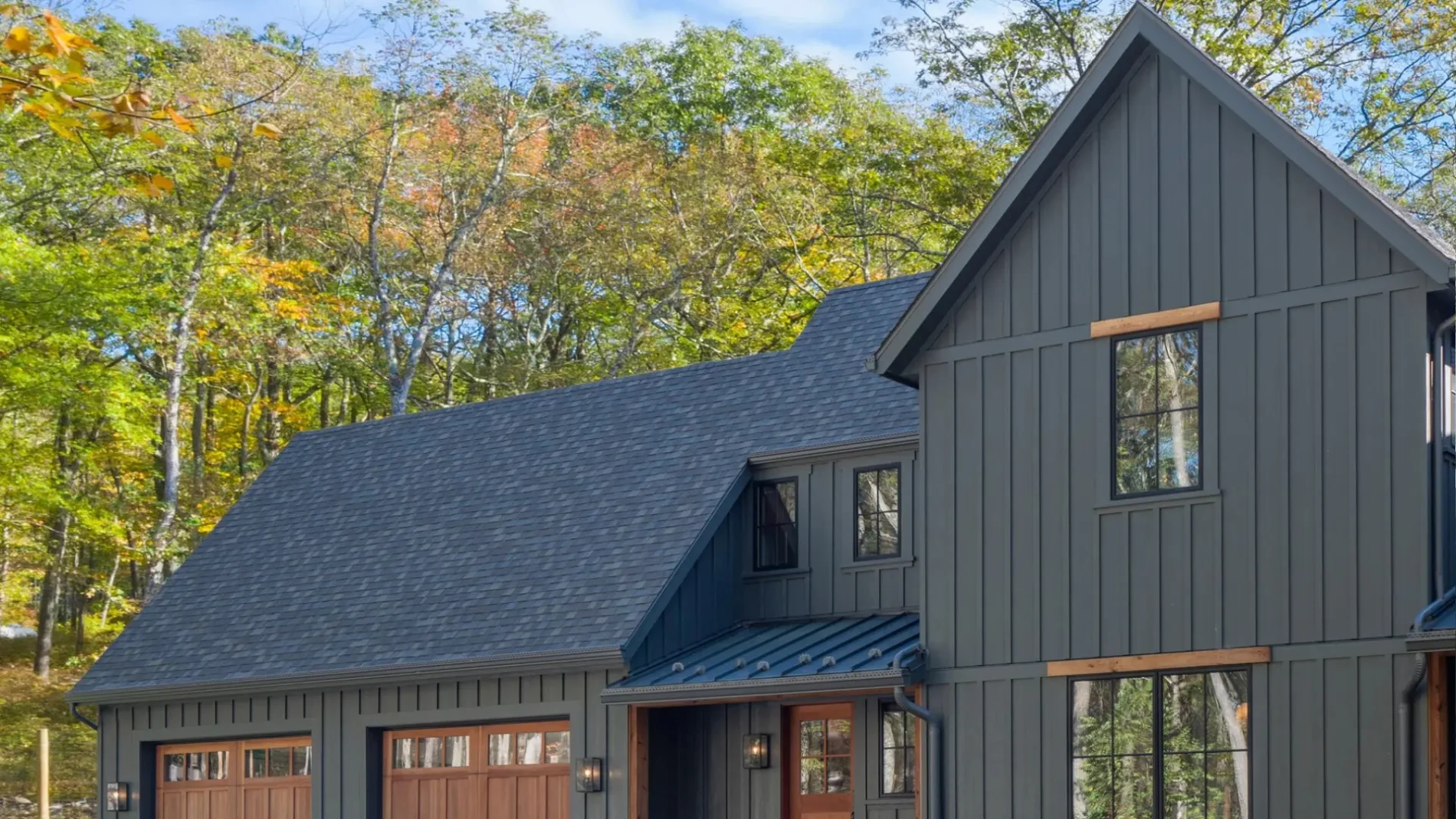 Modern dark gray house with steep shingle roof, vertical board-and-batten siding, and warm wood garage doors, set in front of tall trees.