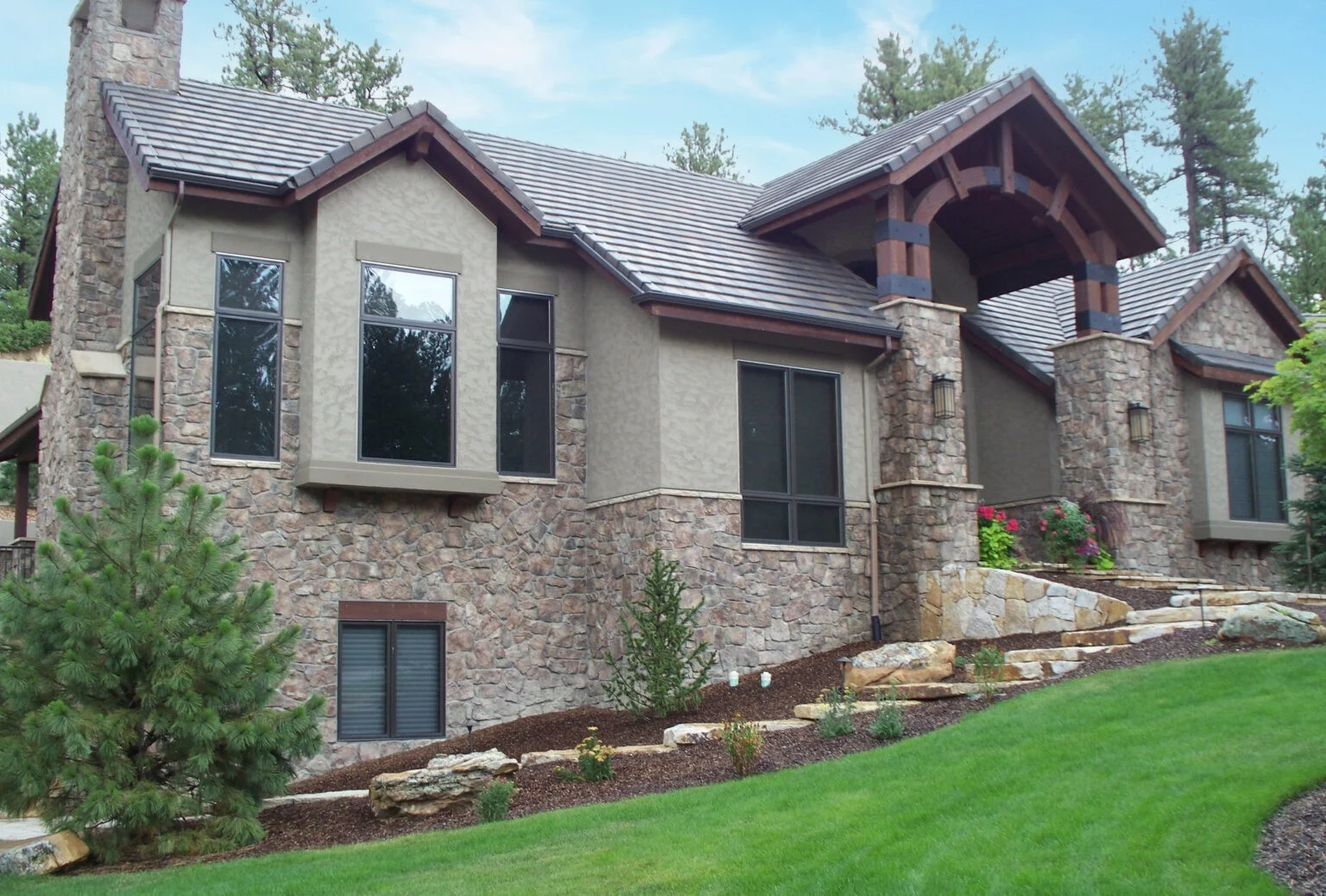 Two-story mountain-style home with stone and stucco exterior, dark-framed windows, and gabled roof, surrounded by manicured shrubs, and rock landscaping.