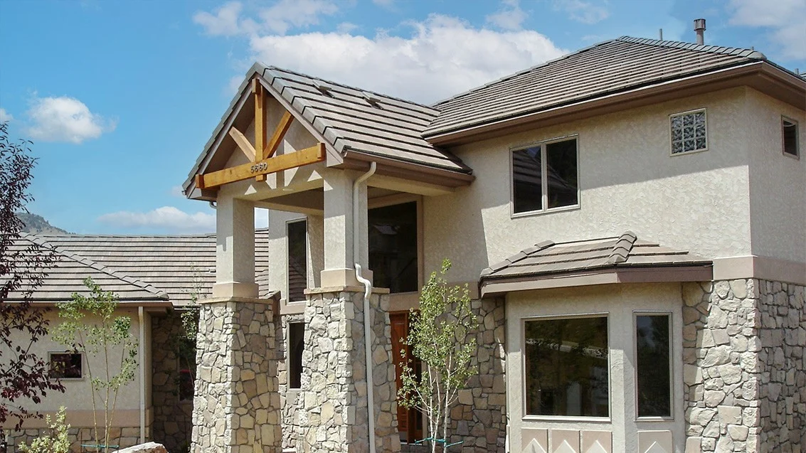 Two-story stucco house with stone accents and a tan tile roof, featuring a gabled front entry supported by stone columns and small trees.