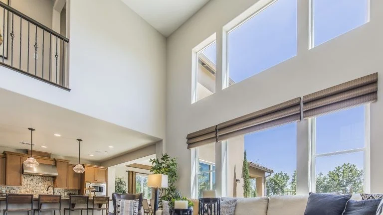 Bright open-concept living room with tall double-height walls, large rectangular windows, neutral furniture, and a view into a modern kitchen.