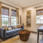 A cozy, sunlit room featuring energy-efficient windows framed by woven shades and floor-length curtains, along with a modern blue sofa and geometric patterned rug.