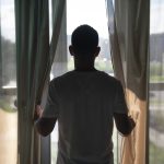 The back of a man holding his curtains open and looking outside through a window.