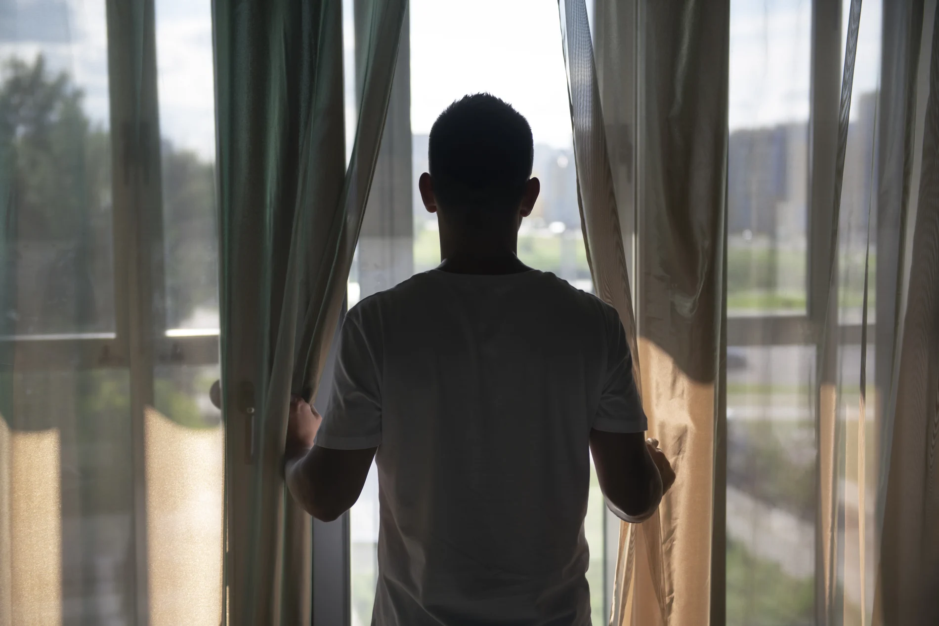 The back of a man holding his curtains open and looking outside through a window.