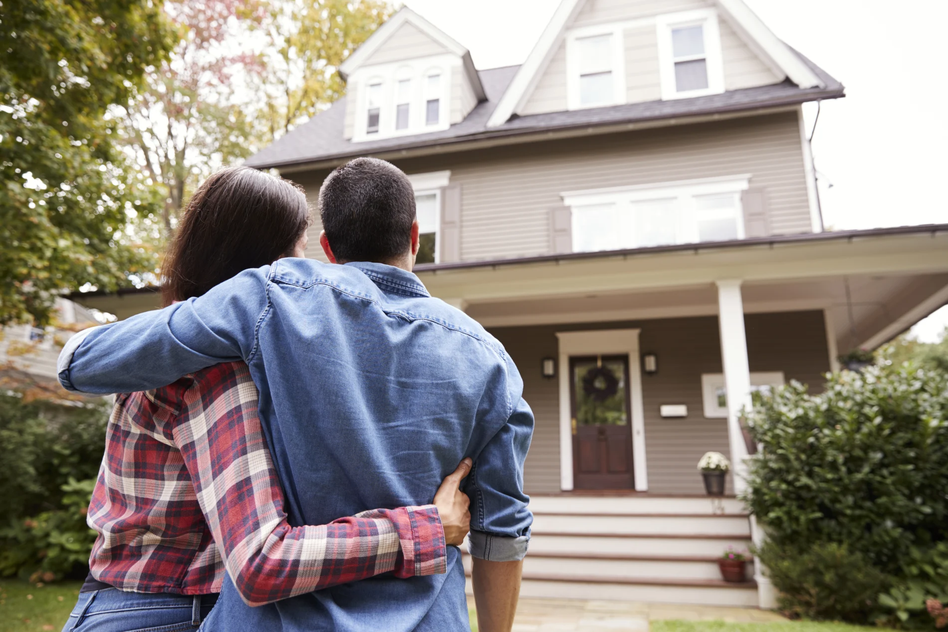 A couple with their arms around each other, looking at their three-story house.