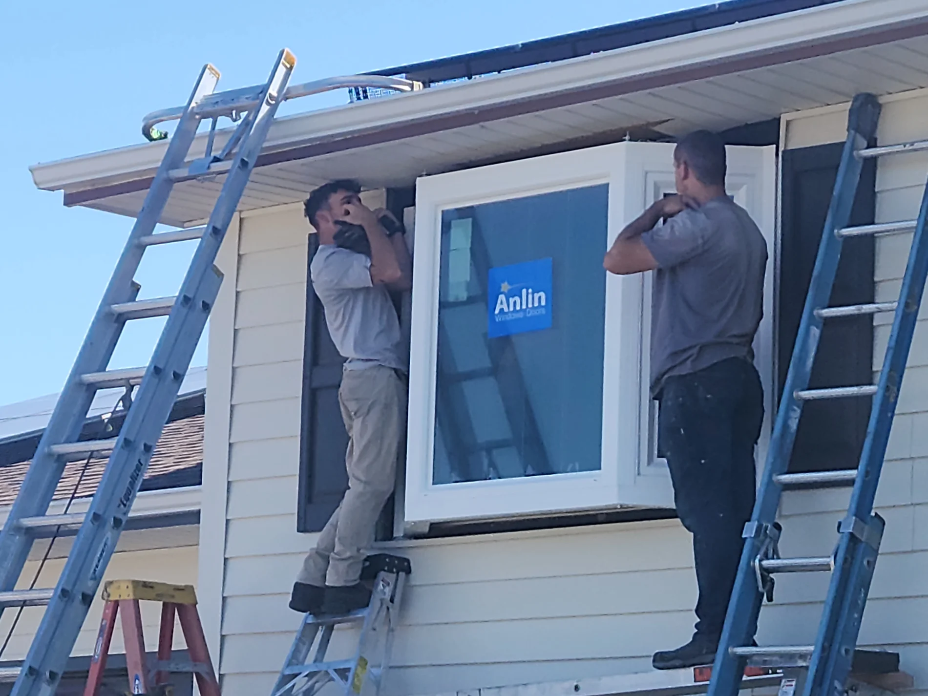 Best Window Installers | Woodland Park, CO | Peakview Windows, Siding, Stucco Two professional window installers stand on ladders to carefully fit a large new white vinyl window into place on the side of a home with light beige siding.