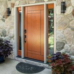 A modern wood-grained front entry door with vertical sidelights and an electronic lock seamlessly integrates with a rustic stone-veneer wall, highlighting a premium door installation flanked by potted coleus plants.