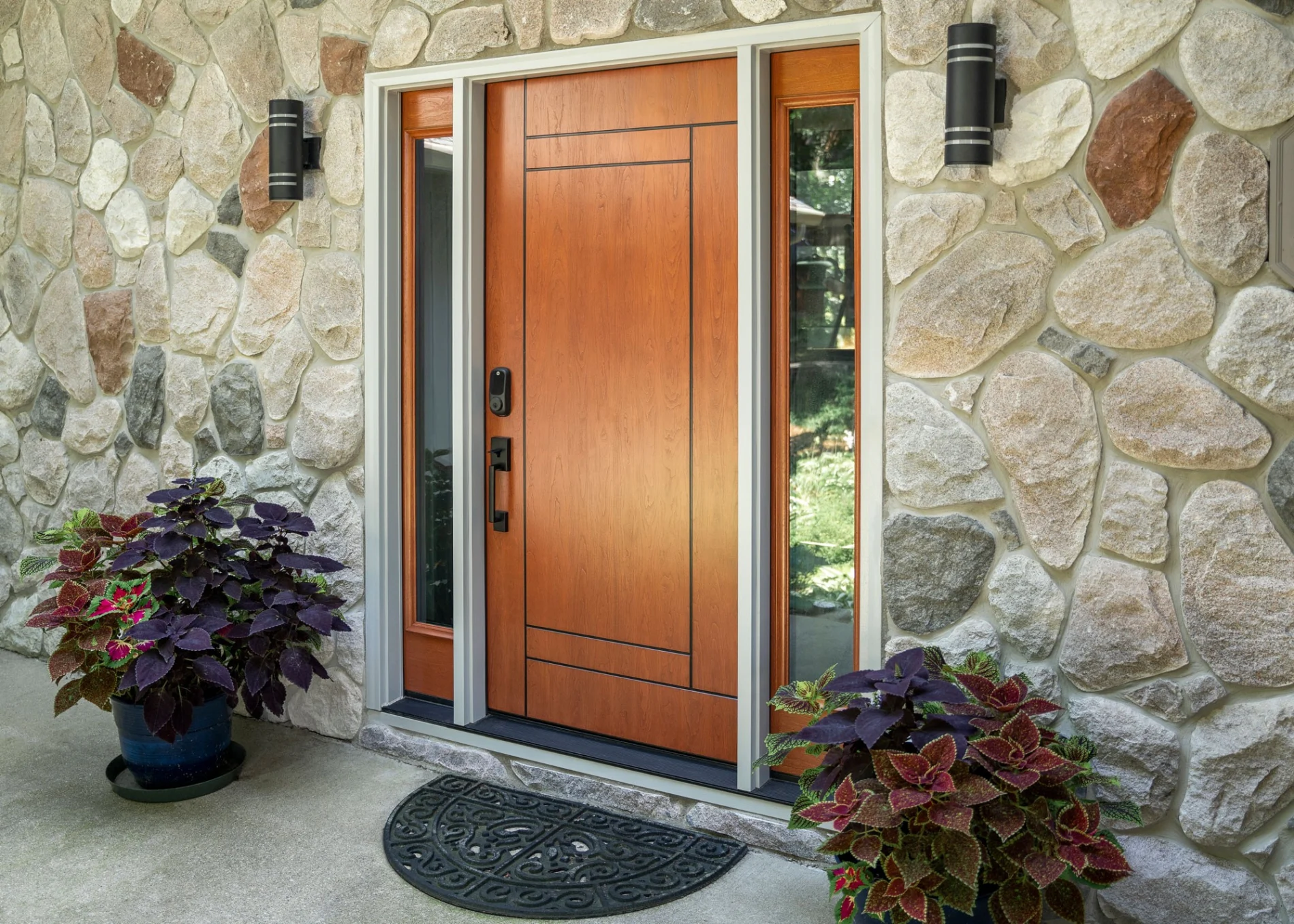 A modern wood-grained front entry door with vertical sidelights and an electronic lock seamlessly integrates with a rustic stone-veneer wall, highlighting a premium door installation flanked by potted coleus plants.