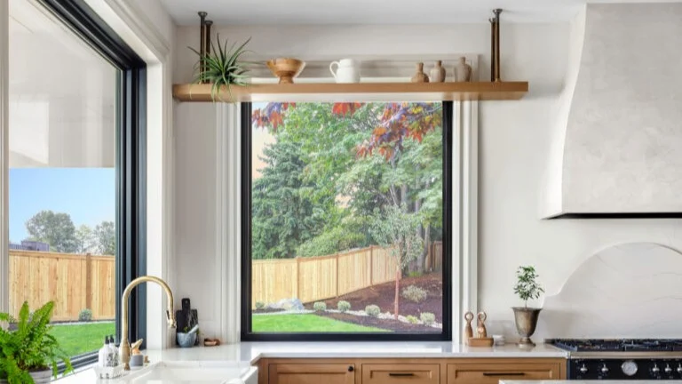 A modern kitchen features a large, black-framed picture window overlooking a landscaped backyard and a wooden fence, showcasing a seamless installation by professional window installers.