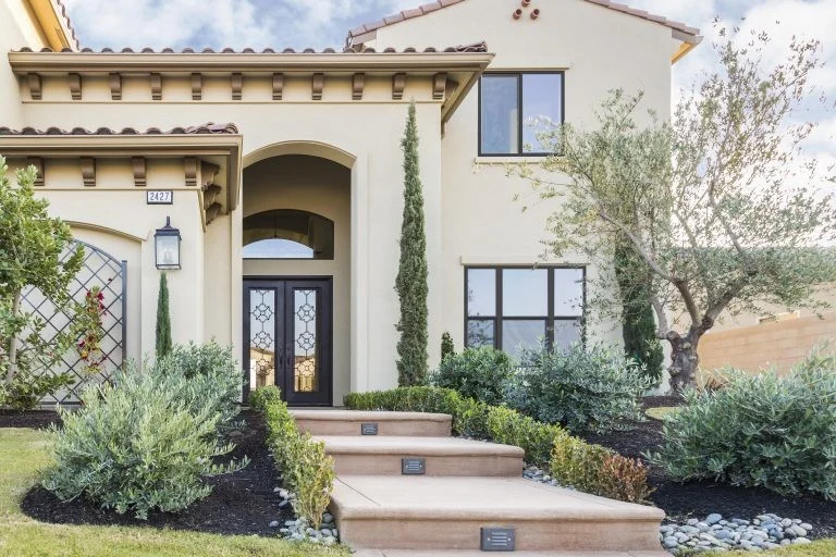 A beautiful Mediterranean-style home features smooth cream exterior walls, an arched entryway with decorative double doors, and a terraced concrete walkway updated by a professional home improvement contractor.