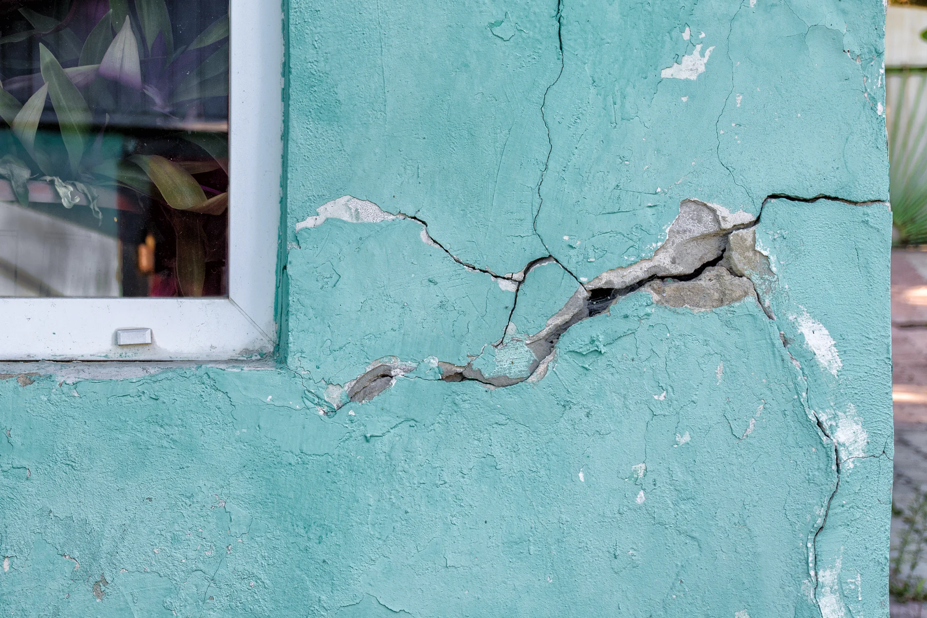 House Wall With A Crack, Destroying The House, The Alarm State Cracks in teal stucco on the side of a house with the corner of a white window