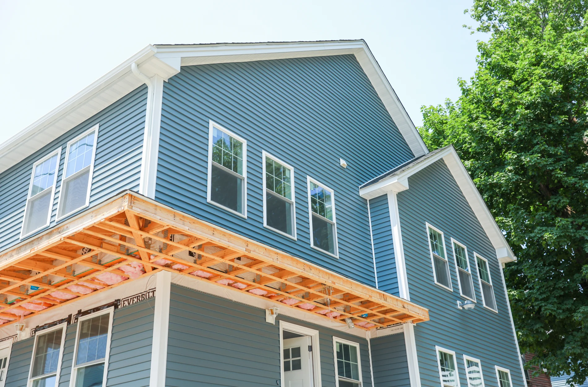 An upward view of a  two-story teal house with white windows, white soffit, and a white door open surrounded by trees under a blue sky.