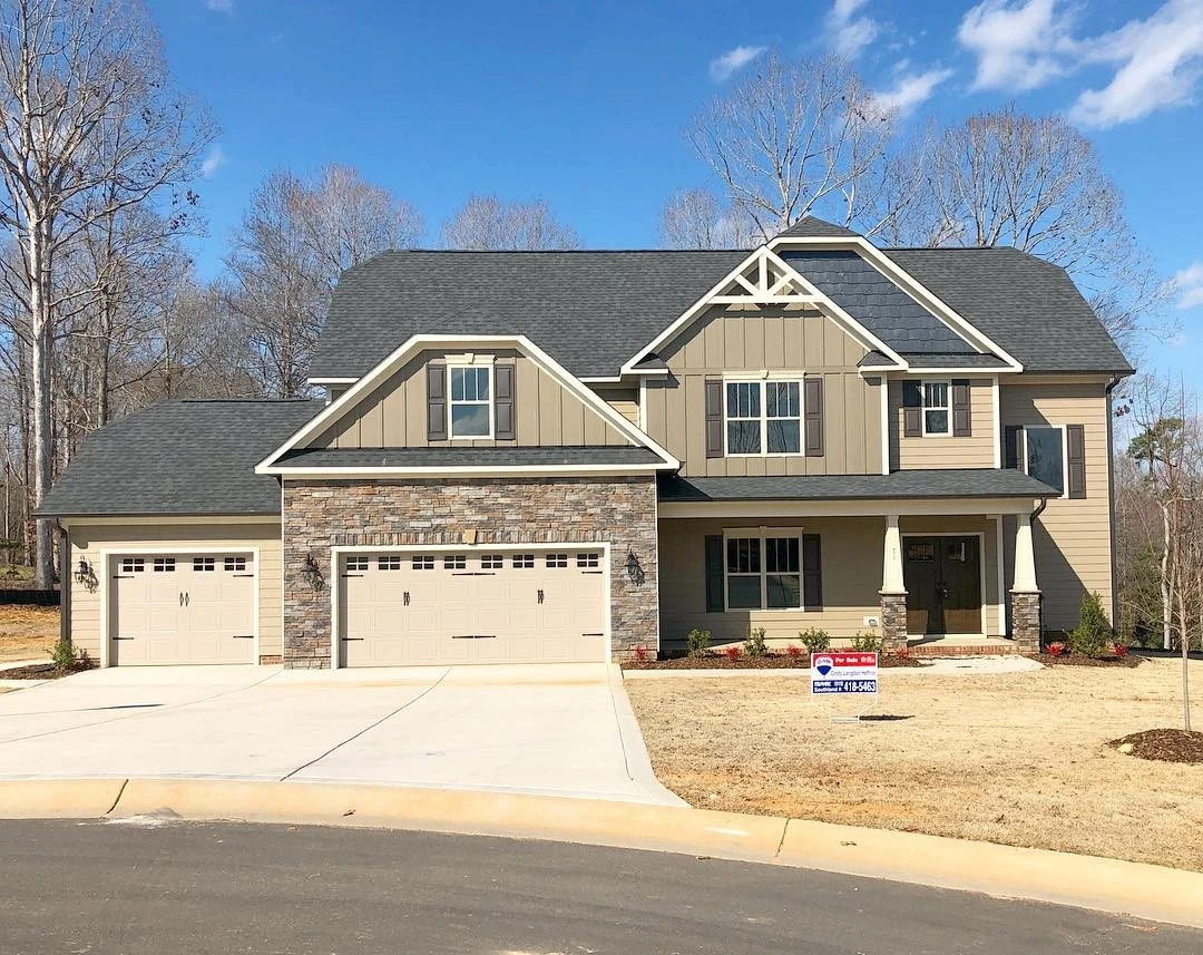 A two-story tan house with stone around one of the two tan garage doors, stone pillars around the wooden front door, white windows with gray shutters, and a shingle roof.