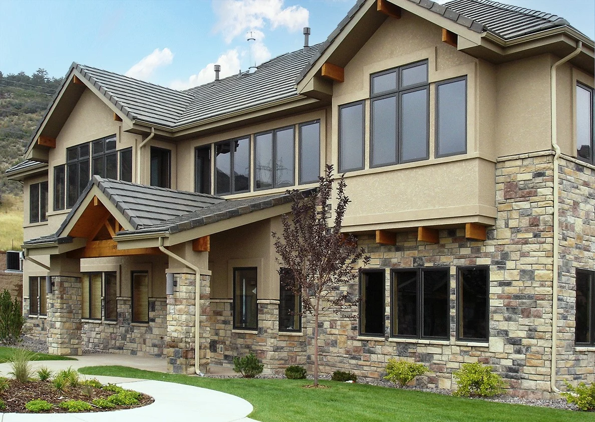 The front of a large two-story residence with the top half having a light brown and textured acrylic stucco siding, and the bottom half covered in Sunset Stone manufactured stone veneer. There are many windows along both stories, a manicured lawn, and simple little bushes in stone landscaping.