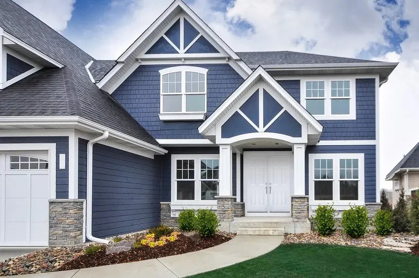A blue two-story house with white double front doors, white windows, white trim, and a shingle roof under a blue sky.